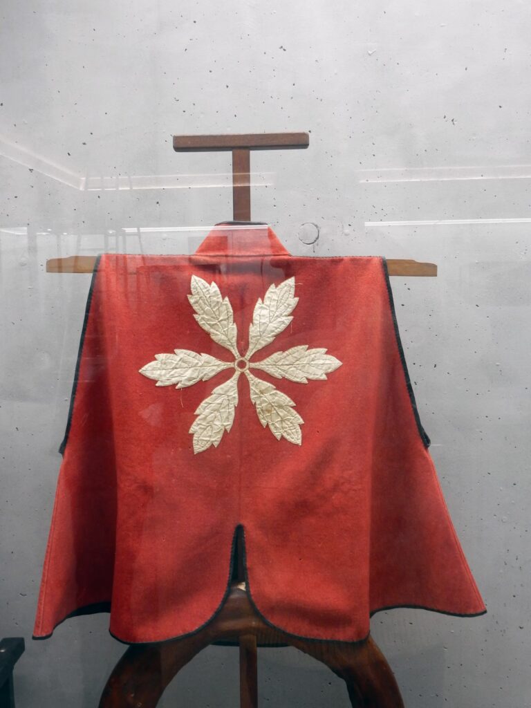 Red woollen jinbaori bearing the Obata clan crest, displayed with a black and red lacquered war fan featuring a rising red sun.