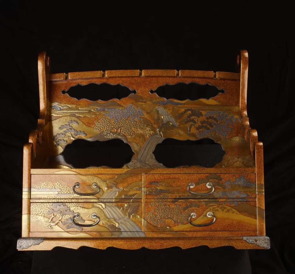 Japanischer Schwertständer (Katana kake) aus der Edo-Zeit (1603–1868) aus der Sammlung des Samurai Museum Berlin. Der Ständer ist mit Gold- und Silberlack verziert und zeigt eine hügelige Landschaft mit Wasserfällen, Bambus, Kiefern und Kirschblüten.