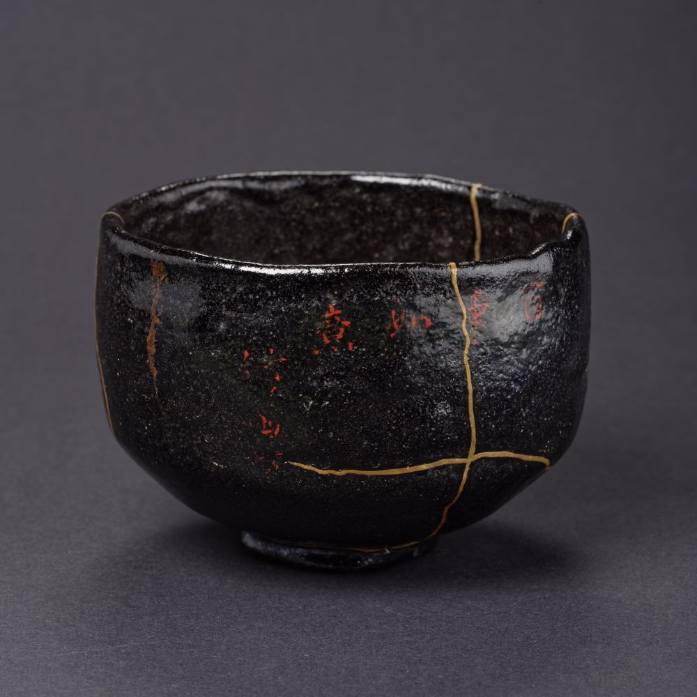 Black Raku tea bowl (kuro-raku) with an asymmetrical hand-shaped form, rough textured surface, gold-filled kintsugi repairs, and traces of red lacquer.