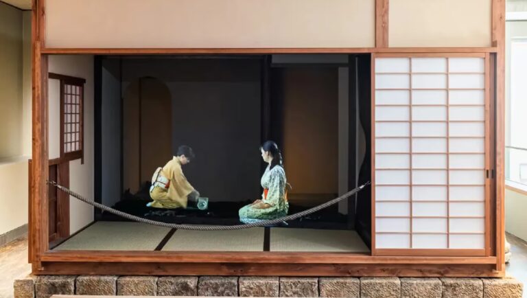Zwei Personen in traditioneller japanischer Kleidung vollziehen eine Teezeremonie in einem Tatami-Raum mit Shoji-Schirmen und Holzrahmen.